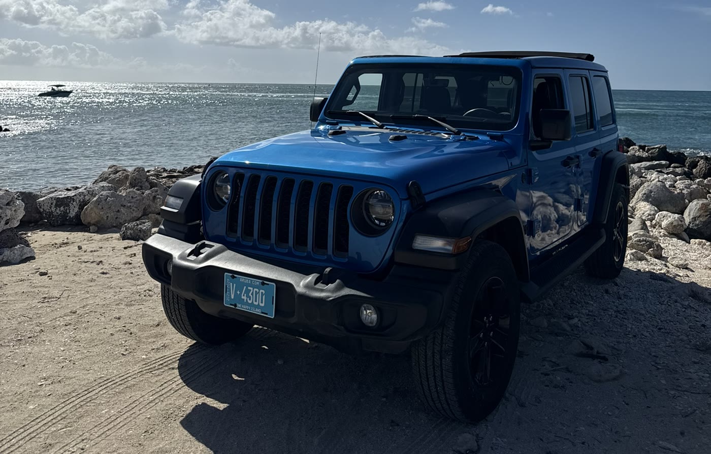 Blue Jeep Wrangler parked on rocky ground by the sea under a blue sky