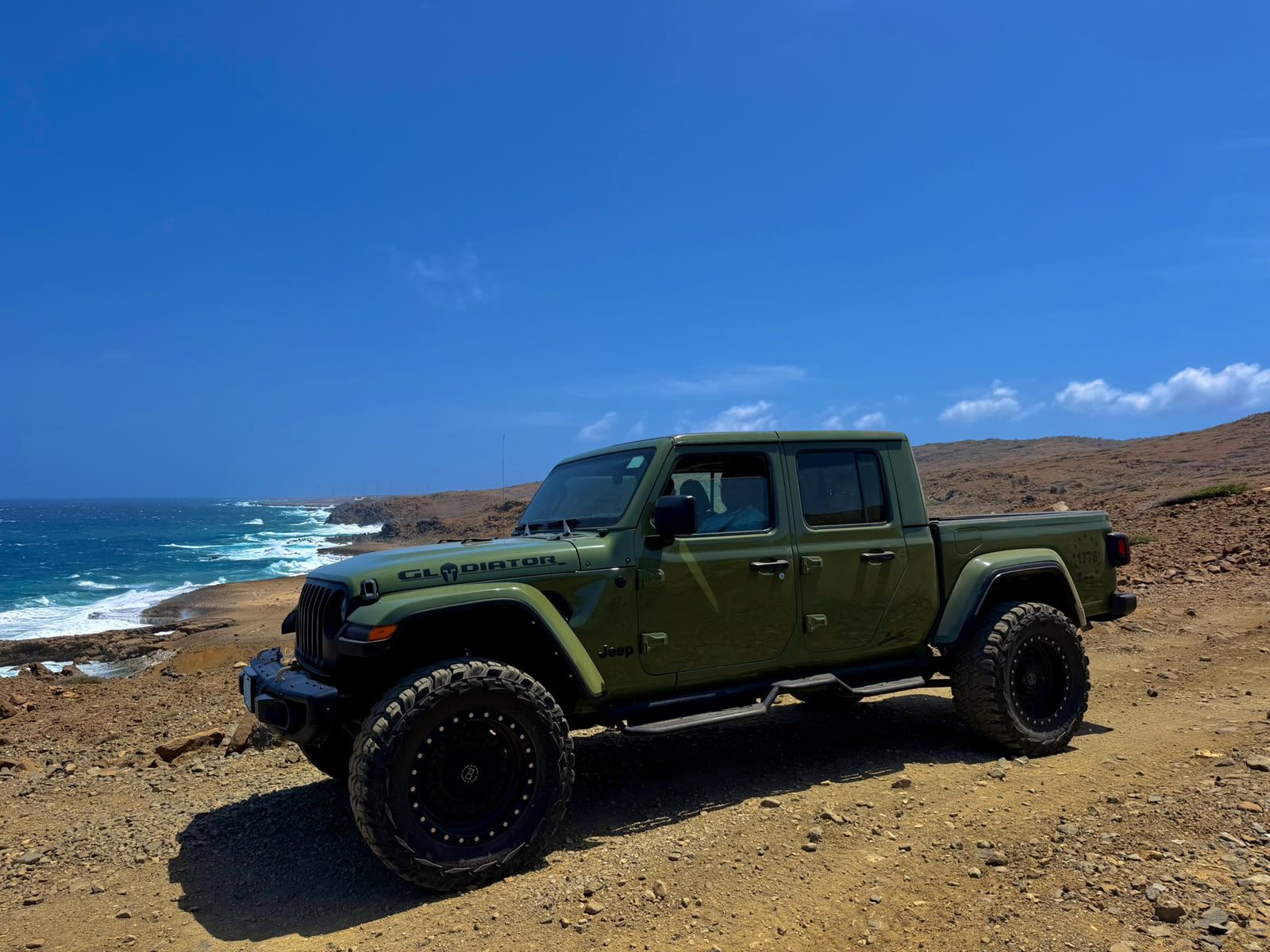 Olive green Jeep Gladiator parked on rocky ground with the sea behind it