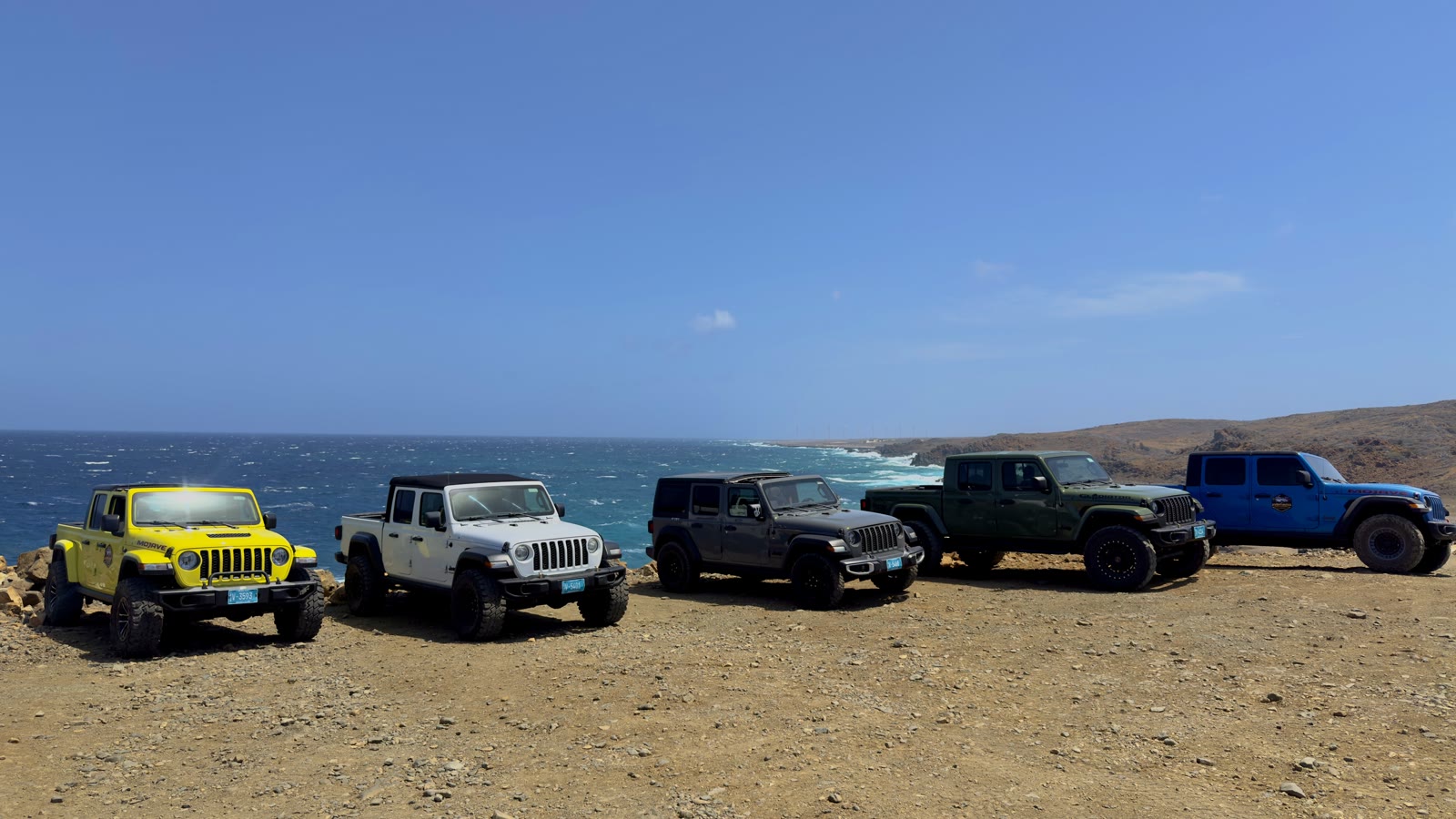 A row of Jeeps in different colours parked on rocky ground above the sea