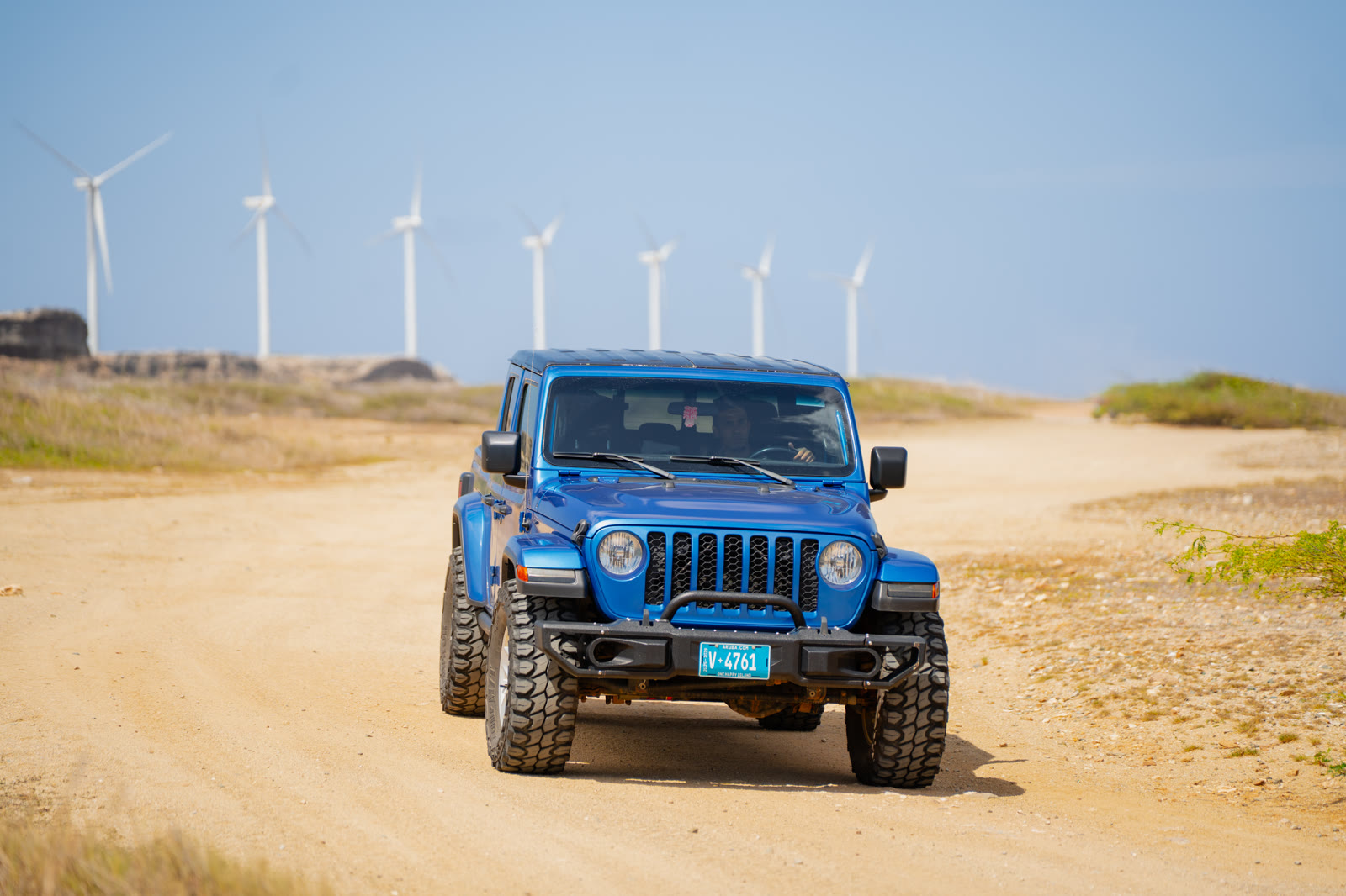 Blue Jeep Wrangler on a sandy road with wind turbines behind it