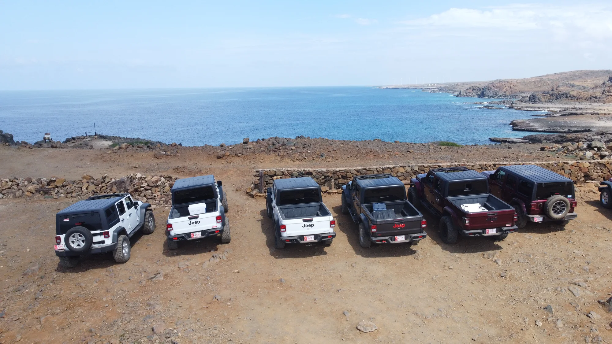 Aerial view of six Aventurova Jeeps parked in a row on rocky ground above the sea
