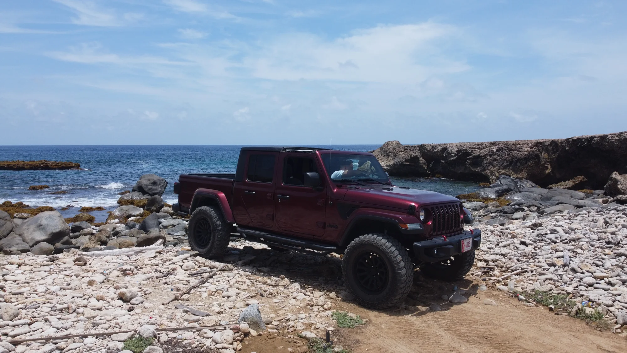 Maroon Aventurova Jeep Gladiator parked on a rocky shoreline beside the sea